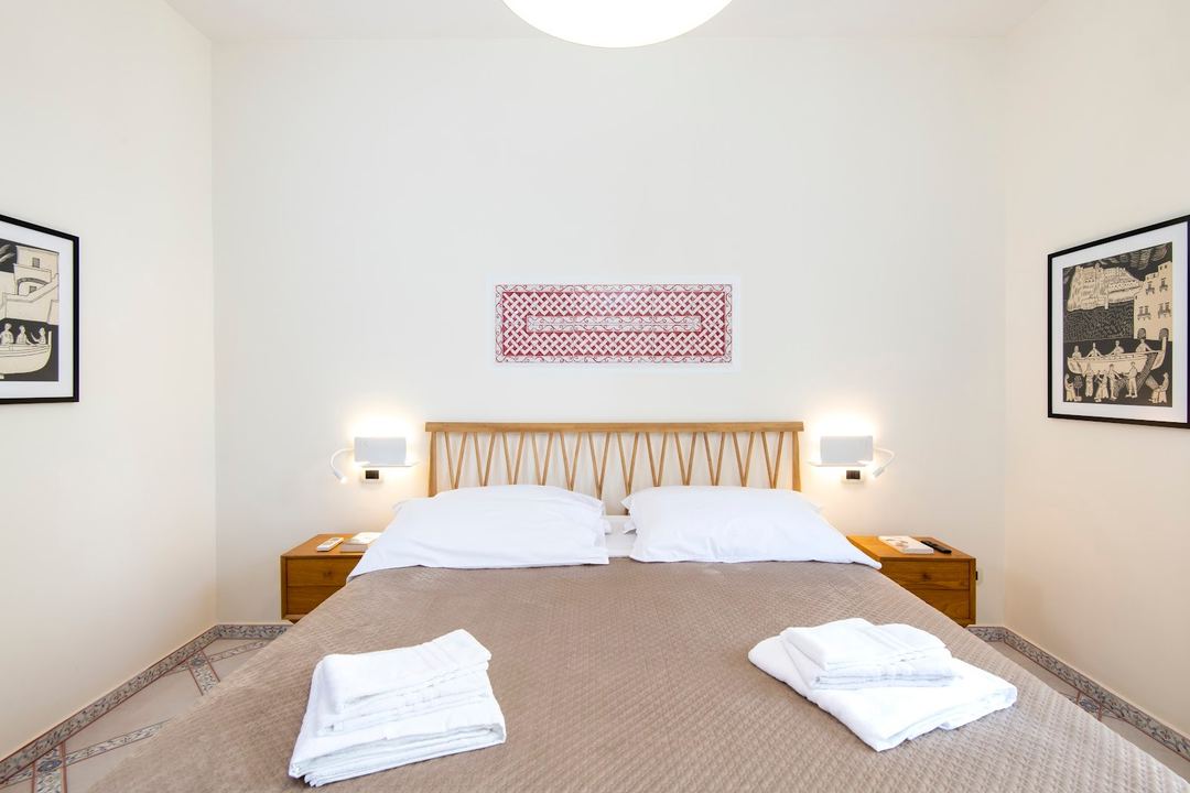 A bright, minimalist bedroom with a wooden bed frame, white pillows, folded towels, and modern wall-mounted lamps.