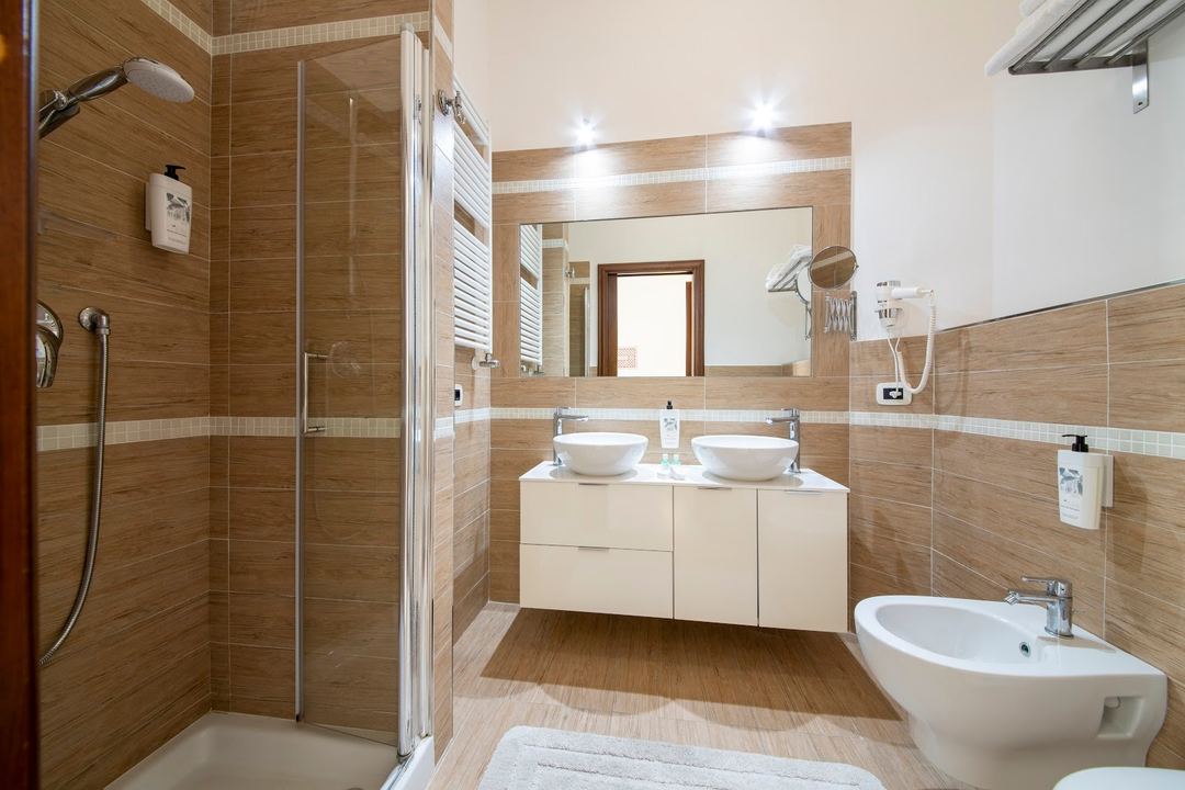 A modern bathroom features dual vessel sinks on a cream vanity, wooden tile accents, a walk-in shower with glass enclosure, and ambient lighting.