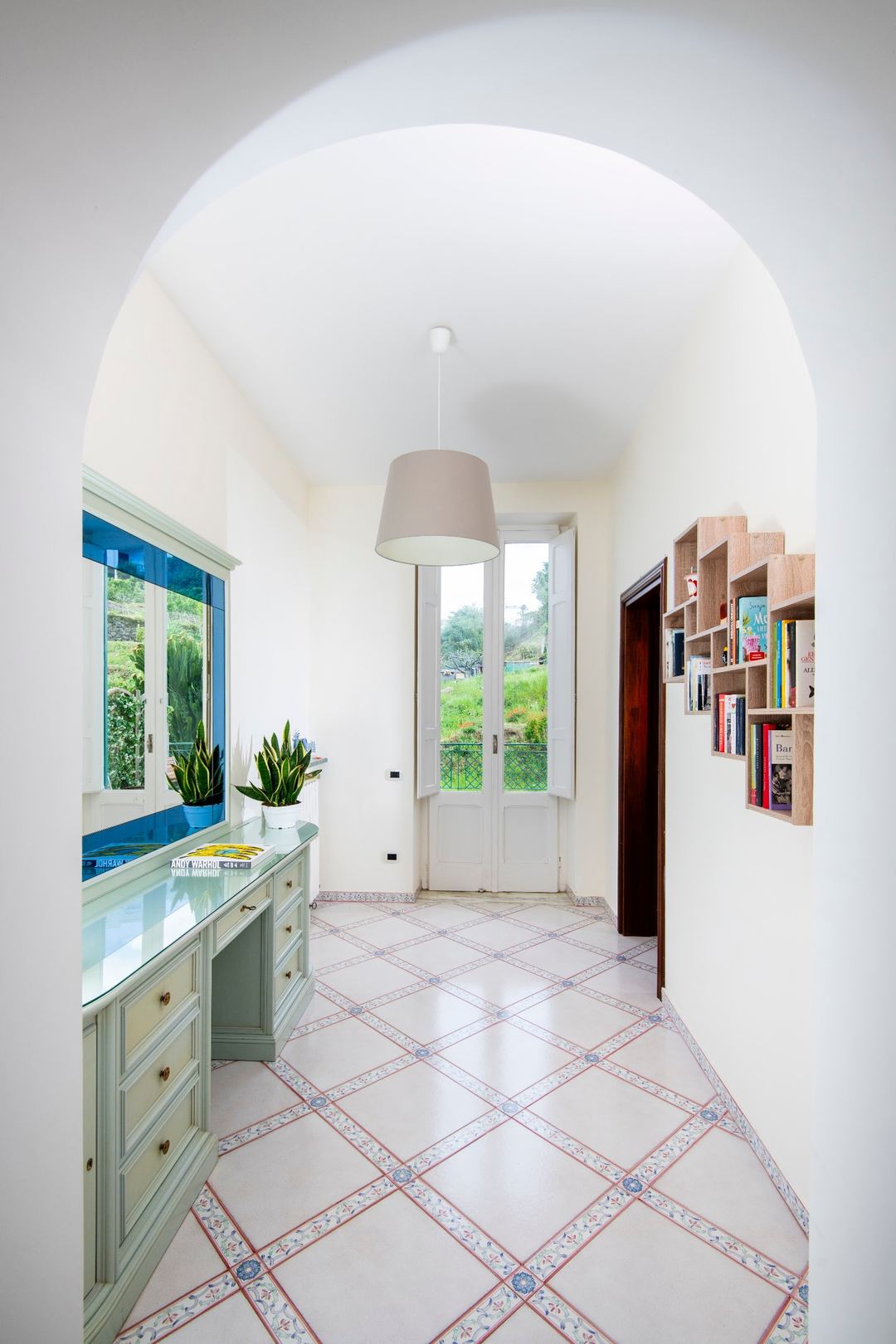 A bright hallway with arched ceilings, decorative terracotta floor tiles, blue-framed windows with garden views, and wooden shelving units.