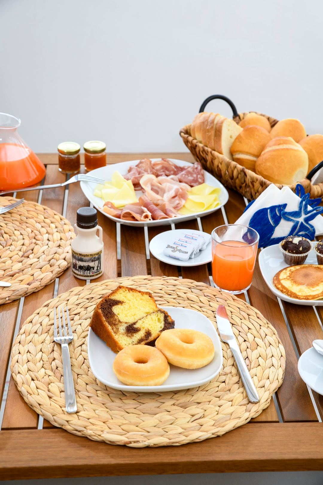 A breakfast table setting featuring donuts, sliced cake, cured meats, cheese, fresh bread rolls, and fresh orange juice.